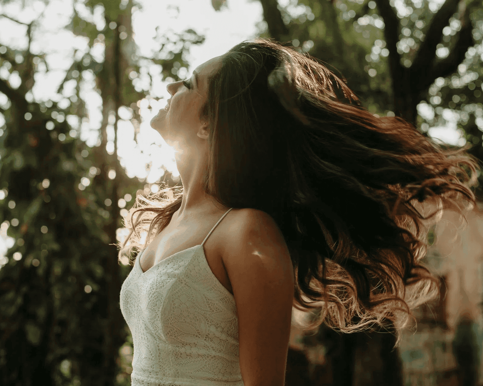 Woman in white dress, hair flowing in sunlight, surrounded by trees.
