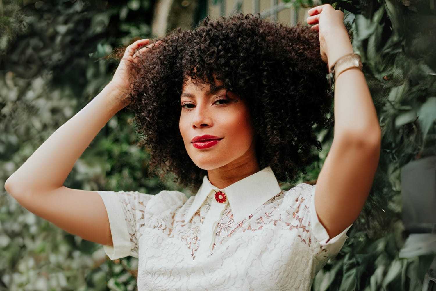 Woman with curly hair in a white lace dress poses outdoors.