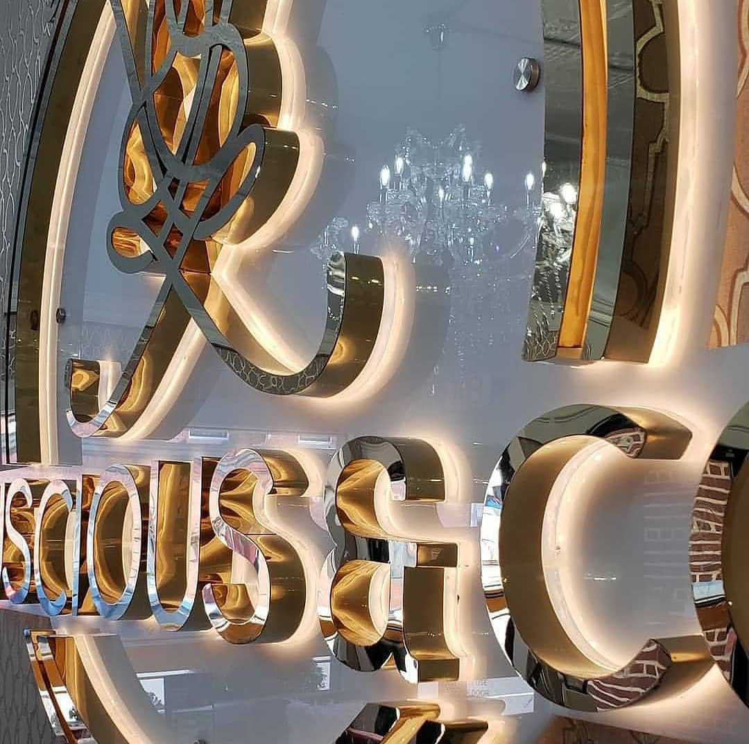 Gold and mirrored sign reflecting a chandelier in a luxury store setting.
