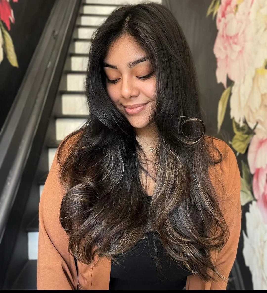 Woman with long hair smiling in front of floral wall and staircase.